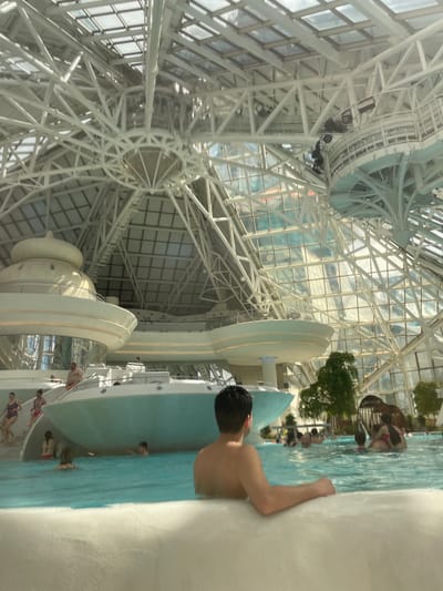 The image shows the Caldea thermal spa in Les Escaldes, Andorra. The spa is located inside a building with a complex structural framework of white metal beams and glass panels. The image is taken from within the pool area, with the camera partially submerged. A man is standing at the edge of the pool, facing towards the center of the water area. Several other people are visible in the pool, some standing and some swimming. There are structures in the middle of the pool painted light blue and white, with light reflecting from the glass ceiling. Some trees and greenery are planted within the spa environment.