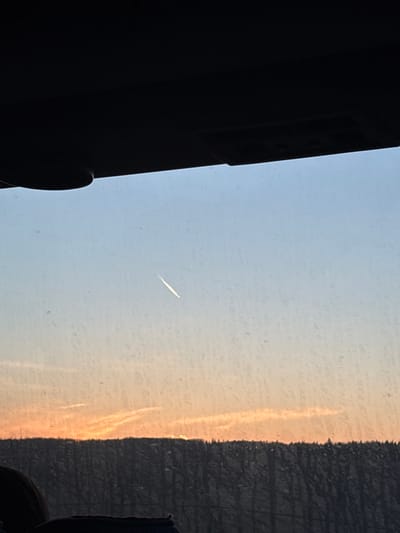 The image shows a view through a window. On the left, there is a blurred dark object, possibly part of the window frame or an object near the window. In the center, there is a pale blue sky with a linear contrail visible. On the right, there is a shadowed object with a textured surface. The light suggests sunset or sunrise. The location (Novoromashkovo, Russia) cannot be confirmed or refuted based on the visual information in this image.