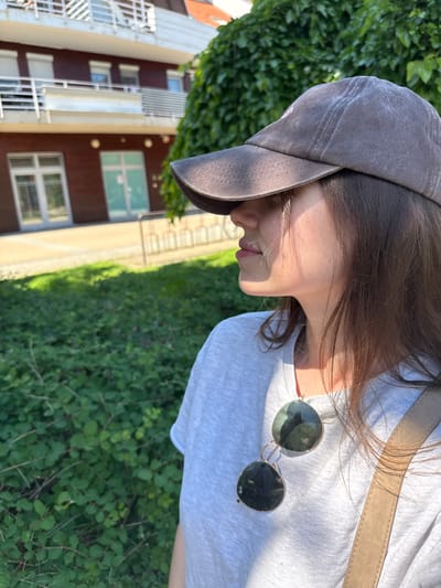 A medium, slightly rotated close-up shot reveals a person wearing a gray baseball cap and a light gray T-shirt. Sunglasses are partially visible, hanging off the T-shirt. A beige strap crosses over the shirt. The individual's profile is visible, showing their face in sunlight. The background features a building with brown and white exterior and greenery, suggesting an outdoor setting. The location appears to be Siófok, Hungary, based on the visible architecture.