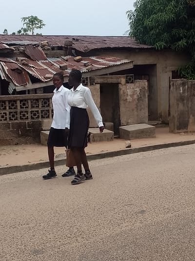 Two women are walking on a paved road. They are dressed in white shirts and black skirts. The building behind them has a partially collapsed corrugated iron roof. The building is likely in a residential area within Biliri, Nigeria. The sky is overcast.