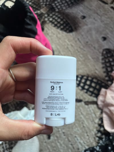 A person's hand holds a white, cylindrical tube of sunscreen. The tube has text printed on it in black and some red lettering. The text mentions "Perfect Balance Sunscreen" and "VERY HIGH PROTECTION". It also includes details about the manufacturer and distributor, as well as a website. The hand holding the tube has a silver ring on one finger. The background is blurred and appears to be an indoor setting with a patterned fabric. The overall impression is of a close-up shot focused on the product and the hand.