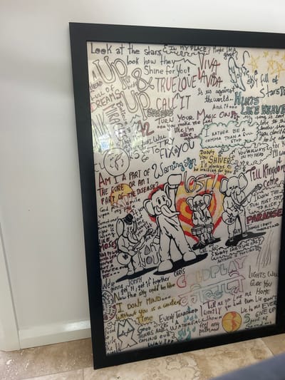 A framed, rectangular artwork with a black frame rests against a plain, light-colored wall and a mottled beige-tiled floor, positioned in Corrimal, Australia. The artwork, executed on a white background, is densely covered with handwritten lyrics and titles from Coldplay songs in various colors, primarily black, red, and blue. Prominent song titles include "UP&UP," "VIVA LA VIDA," "FIX YOU," "LOST!," and "PARADISE." In the central-lower section, three cartoon elephants are illustrated playing musical instruments: one with a guitar, one with drums, and another standing with an arm raised. Additional graphic elements include stars, wing outlines, and abstract shapes. Reflections are visible on the artwork's protective covering. A white baseboard is present along the wall-to-floor junction.