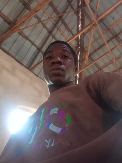A single individual, a male with dark skin and short dark hair, is depicted in a low-angle, head-and-upper-torso shot, looking directly into the camera. He wears a brown garment with multicolored abstract embroidery on the chest, including green, purple, blue, white, and orange elements, and partially visible lighter-colored text below. The background features an indoor environment with an exposed ceiling structure consisting of wooden rafters supporting corrugated metal or panel roofing. A light-colored wall is visible on the left. A bright, localized light source creates glare in the lower-left portion of the image. The scene is located in the city of Isinyi-Nando, Nigeria.
