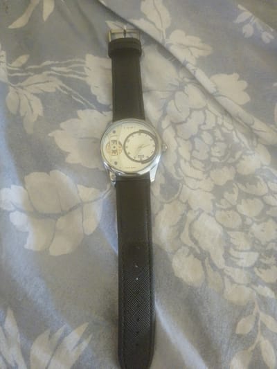 A black wrist watch is laid out on a light-colored fabric with a floral pattern. The watch has a silver casing and a white face with black markings and hands. The strap is black and appears to be made of a textured material. The fabric background is a light blue-grey with a pattern of stylized white flowers and leaves. The scene is viewed from directly above. There are no people present in the image.