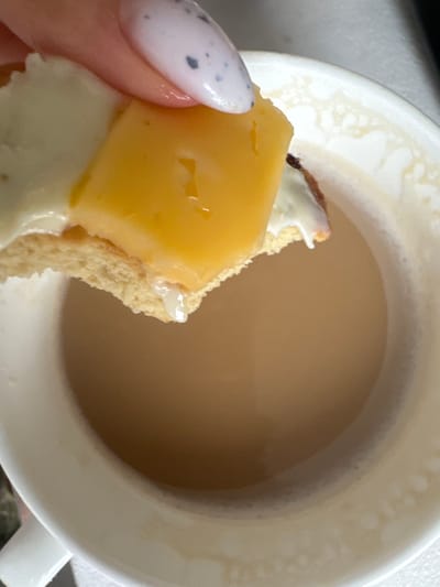 The scene depicts a close-up view in Gribovo, Russia, showing a human hand interacting with food and a beverage. A hand with a manicured fingernail, painted light with dark speckles, holds a piece of a light-colored baked good. This baked good is topped with a white creamy substance and a rectangular slice of yellow cheese. A droplet of liquid hangs from the bottom edge of the baked good. Below this, a white ceramic mug, displaying residue marks on its inner rim, contains a light brown, opaque liquid. The baked good is positioned directly above the liquid, appearing to be dipped or about to be dipped into it.