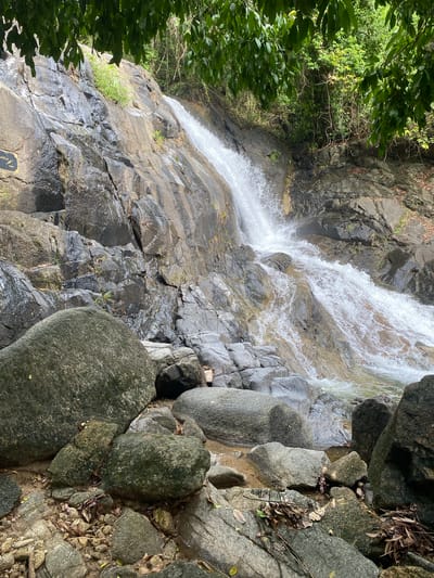 The image shows a waterfall cascading over a rocky cliff face. The water is white and foamy, contrasting with the dark grey and black rocks. Patches of green vegetation cling to the rocks on the left side of the image. The scene is partially obscured by foliage in the upper left corner. Debris is scattered on the rocks. The image appears to be shot in Thai Mueang, Thailand.