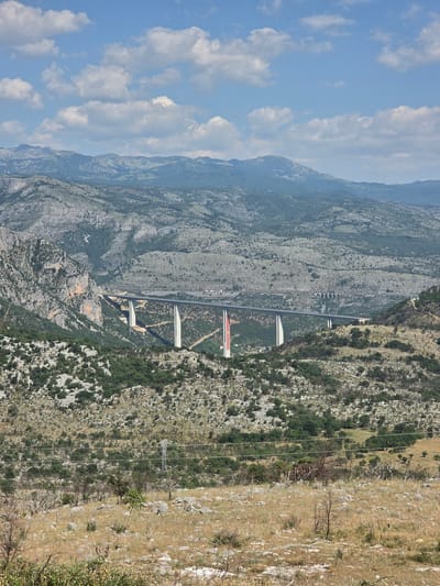 The image captures a sweeping, vertical panorama of a mountainous landscape in Blizna, Montenegro. The scene is dominated by rugged, grey mountainsides covered in patches of green vegetation, primarily shrubs and trees. A striking modern bridge, featuring multiple concrete pillars and a long, elevated roadway, cuts across the valley, connecting different parts of the mountainous terrain. The bridge appears to be part of a highway system, with at least two distinct sections visible, one with a lighter-colored roadway and another with a darker surface.

In the foreground, the right side of the image shows a drier, more arid slope covered in dry grass, rocks, and scattered bushes. A series of power lines stretch across this part of the landscape, supported by poles, indicating infrastructure in the area. The sky above is a vibrant blue, dotted with fluffy white clouds, suggesting a clear and pleasant day.

The time of day appears to be daytime, likely mid-morning or afternoon, given the bright sunlight and the shadows cast by the bridge and the terrain. The weather seems fair and sunny. There is no visible text in the image. The overall impression is one of a grand natural landscape with a significant modern engineering feat integrated into it. The scale of the mountains and the bridge suggests a remote and impressive location.