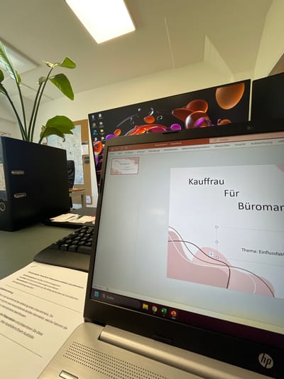 This media file captures a desk setup with a laptop and an external monitor, suggesting a work or study environment. The laptop is open, displaying a presentation slide with the title "Kauffrau Für Büromanagement" (Trainee for Office Management) and the subtitle "Thema: Einflussfaktoren" (Topic: Influencing Factors). The presentation appears to be in German, as indicated by the text.
The scene is likely indoors, possibly an office or a home office, given the presence of office supplies like a black binder and a corkboard with what looks like a map pinned to it. A plant with large green leaves adds a touch of nature to the workspace. The lighting suggests it might be daytime, with a bright light source visible on the ceiling. The overall mood conveyed is one of productivity and learning. The image is taken from a slightly angled perspective, allowing a view of both the laptop screen and the external monitor, which shows a colorful abstract background. A part of the laptop's keyboard is also visible. The HP logo is discernible on the laptop. The context of Morsbach, Germany, places this scene within a specific geographic location, though no explicit landmarks are visible.
