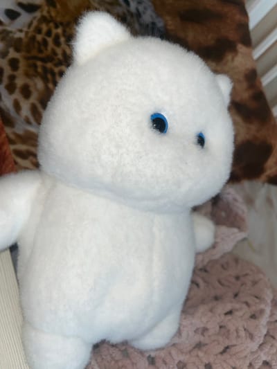 A white plush toy bear with bright blue eyes is lying on a bed. The bear is positioned on its side, with its head slightly turned to the left. To the left of the bear, a blanket with a brown and black leopard print is visible. Behind the bear, a pink knitted blanket is partially seen, along with the edge of a white wooden crib or bed. The lighting suggests it is daytime, with soft, diffused light. There is no text visible in the image. The scene appears to be a comfortable and safe resting place, possibly a nursery or a child's bedroom.