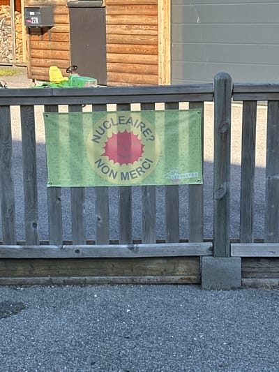 The image shows a banner attached to a wooden fence. The banner is green with a yellow circle in the center. Inside the circle, it reads "NUCLEAIRE? NON MERCI!" in French, meaning "Nuclear? No Thanks!". There is also a red sun-like symbol with a smiley face inside the circle. In the lower right corner of the banner, there is a logo that says "LES VERT-E-S", which refers to the Green Party in Switzerland.
In the background, there is a wooden building with a mailbox marked "27A". A toy lawnmower with a yellow seat is visible in front of the building. The fence is made of vertical wooden planks. The ground in front of the fence is covered with asphalt or gravel. The scene appears to be outdoors, likely in a residential or semi-rural area. The time of day seems to be daytime, with some shadows indicating sunlight. The city of Le Sentier, Switzerland is a commune in the canton of Vaud, known for its watchmaking industry. The overall setting suggests a peaceful, possibly domestic environment, with the banner conveying a political or environmental message against nuclear energy.