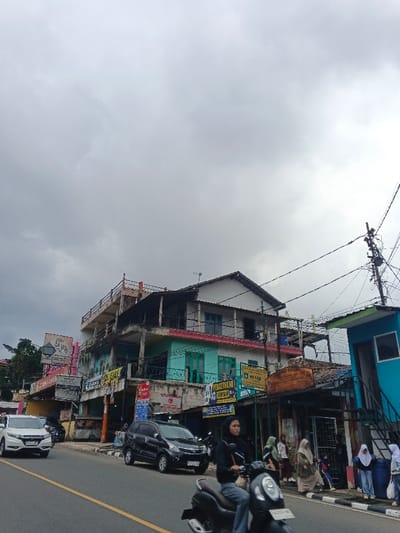 The scene is located in Palayan, Philippines, depicting a busy street under an overcast, gray sky. The foreground shows an asphalt road with a solid yellow dividing line. In the immediate foreground, a person wearing a black long-sleeved shirt and a black hijab is riding a dark gray/silver scooter-type motorcycle, moving from right to left.
To the left, a white Honda CR-V SUV is traveling in the opposite direction, while a black SUV, possibly a Toyota Avanza, is visible parked or moving slowly on the right side of the road, behind the scooter.
On the right side of the street, a row of multi-story buildings lines the road. A prominent building features a white upper section with a gabled roof and teal-colored window frames on the lower level, accented by a red-painted balcony railing on the second floor. Various commercial signs are affixed to the buildings, displaying text such as "PERCETAKAN FRESH MILK" and "SELAMAT DATANG." Further right, a bright blue structure with external metal stairs is visible, and overhead, numerous entangled electrical wires and utility poles are present.
A group of approximately 5-7 pedestrians, including several individuals wearing hijabs and at least one child in a blue and yellow outfit, are seen on the sidewalk alongside the buildings. Sparse green foliage is visible on the far left, behind the buildings.
