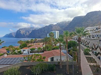 The image depicts a scenic view of a coastal town in Santiago del Teide, Spain, with the iconic Acantilados de los Gigantes (Cliffs of the Giants) serving as a dramatic backdrop. The foreground features a residential area with white buildings, terracotta roofs, and lush green vegetation, including palm trees and cacti. Solar panels are visible on one of the rooftops, indicating a focus on sustainable energy. A paved walkway winds through the community, bordered by carefully landscaped gardens. In the mid-ground, a modern white hotel building stands tall, surrounded by palm trees. The background is dominated by the imposing cliffs, rising majestically from the deep blue sea. The sky is partly cloudy, with patches of blue sky peeking through. The overall scene conveys a sense of tranquility and natural beauty, with a blend of modern architecture and a Mediterranean coastal landscape.