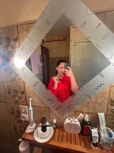 The image captures a female individual taking a mirror selfie inside a bathroom located in Ancenis, France. The subject, visible from the chest up, is reflected in a diamond-shaped mirror framed with frosted geometric patterns. She is wearing a red, collared garment and holds a dark-colored smartphone in her right hand to capture the reflection. A ring is present on her right ring finger. Her dark hair is pulled back, and her left hand is positioned near her shoulder.
The reflected background behind the individual shows a light brown tiled wall with floral patterns on the left, a wooden door, and a white paneled door or cabinet on the right. A red horizontal bar is mounted on the wall. A light source on the far left creates a bright glare in the mirror.
The foreground displays a wooden slatted countertop holding various personal care items. From left to right: a light blue and white electric toothbrush on its charging base, plugged into a white double electrical outlet; a shaving brush with a dark handle in a white stand; a small dark bottle; a white ceramic toothbrush holder decorated with two cartoon figures, containing multiple toothbrushes; a green spray bottle; and a tube labeled "DENTIFRICE BIO" (organic toothpaste) in a white container. The wall directly behind these items is tiled with a textured, light brown surface.