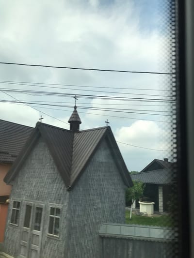 The image depicts an outdoor scene in Vicovu de Sus, Romania, dominated by a small, grey-sided building with a dark brown metal roof. The building's exterior is constructed from uniform, horizontally arranged grey wooden shingles or siding. A central, dark brown, conical spire, topped with a cross, rises from the roof's peak. Two additional crosses are positioned on the roof ridges to the left and right of the spire. On the visible facade of this building, a central double door, each leaf comprising two rectangular panels, is flanked by two single-pane windows with white frames.

Behind the grey-sided building, a portion of a larger residential building with a brown tiled roof is visible to the left. Further to the right and in the background, another residential structure with a dark roof and white-columned elements is partially visible amidst green foliage. In the yard of this background house, a white, cylindrical well structure, topped with a dark apparatus, is present on a patch of green grass.

The sky is overcast with a mix of light grey clouds and patches of brighter white cloud formations. Multiple black utility lines, varying in thickness, traverse horizontally across the upper third of the frame. The extreme right edge of the image displays a dark, textured vertical element, suggesting the photograph was taken from behind a patterned screen or a vehicle window frame. No people are visible in the scene.