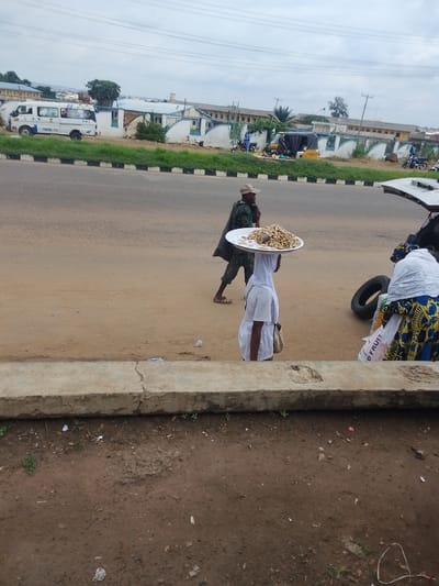 The image shows a street scene in Oja Oba, Oko Erin, Nigeria. The sky is overcast, suggesting it might be daytime with cloudy weather.
In the foreground, a person wearing a white headscarf and white top carries a large tray of peanuts on their head. They are walking towards the right side of the image. Behind them, another person in a camouflage-patterned outfit and a cap is also walking, moving away from the camera.
To the right of the person with the tray, a car's trunk is open, and a person in a colorful patterned dress and a white headscarf is interacting with the contents of the trunk. A large black tire is visible near the trunk. A bag with the words "D FRUIT" printed on it is also visible.
In the background, there is a road with vehicles, including a white van with blue and red markings. Several buildings with white walls and blue trim line the street behind a fence. A few people are seen near some motorcycles and a yellow tricycle. The overall scene depicts a busy outdoor marketplace or roadside area.