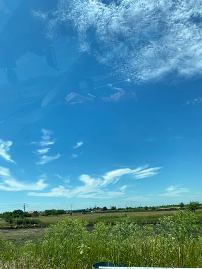 The image shows a view from inside a vehicle looking out through the windshield. The sky is a vibrant blue with scattered wispy white clouds. In the reflection on the windshield, parts of the car's interior are visible, including a rear-view mirror and a person's hands on the steering wheel.
Beyond the windshield, a rural landscape unfolds. A grassy field stretches towards a row of houses with red-tiled roofs and some trees. Power lines with tall poles are also visible in the middle ground. The foreground consists of lush green vegetation, including tall grasses and some flowering plants.
The scene appears to be in a suburban or rural area, possibly on the outskirts of Zrenjanin, Serbia, given the context provided. The lighting suggests it is daytime, likely under clear or partly cloudy skies. There are no overt signs of activity or specific events occurring in the image, beyond the implied presence of the vehicle and its occupants.