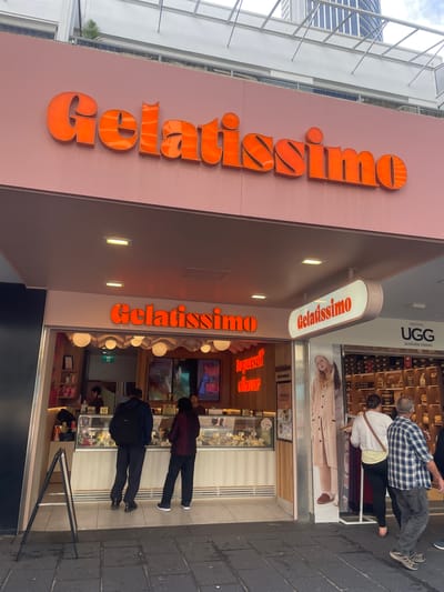 In Surfers Paradise, Australia, a storefront for "Gelatissimo" is prominently displayed. The name "Gelatissimo" is emblazoned in large, vibrant orange, three-dimensional lettering across a pale pink facade above the entrance. Beneath this, a smaller, illuminated oval sign also displays "Gelatissimo" in orange lettering. The shop appears to be a gelato parlor with a display case showcasing various flavors of gelato. Inside, staff members are visible behind the counter. Customers are seen in front of the counter, some browsing and making selections. To the right of the gelato shop, there is a neighboring store with a sign for "UGG" and displays of footwear. The street in front of the shops is paved with dark grey cobblestones. Two individuals, one wearing a blue and white checkered shirt and another in a white top and black pants, are walking away from the viewer towards the right side of the frame, appearing to be customers. A third person, mostly obscured, is walking ahead of them. A large promotional display featuring a woman in a fuzzy robe stands near the entrance of the gelato shop. The overall environment suggests a commercial street scene in a popular tourist area.