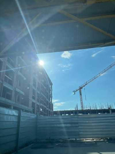 A construction site in Plovdiv, Bulgaria, is captured on a clear, sunny day. The image is framed at the top by the underside of a light blue-grey corrugated metal canopy or roof structure, supported by yellow-painted metal beams, with prominent sun flare radiating from the upper left quadrant. To the left, a multi-story building is under construction, featuring exposed reddish-brown ceramic blocks forming walls and grey concrete slabs for floors and balconies. The sun's glare partially obscures the top of this building. To the right, an orange tower crane, with "CAEL" visible on its mast, extends its long arm diagonally across the bright blue sky, which contains scattered white clouds. Below the crane, another section of the construction site shows concrete foundations or floor slabs with numerous vertical steel rebars protruding upwards. A light grey, horizontally corrugated metal fence acts as a perimeter barrier in the lower portion of the image, separating the construction area from the immediate foreground, which shows a concrete or dirt surface. No individuals are visible in the scene.