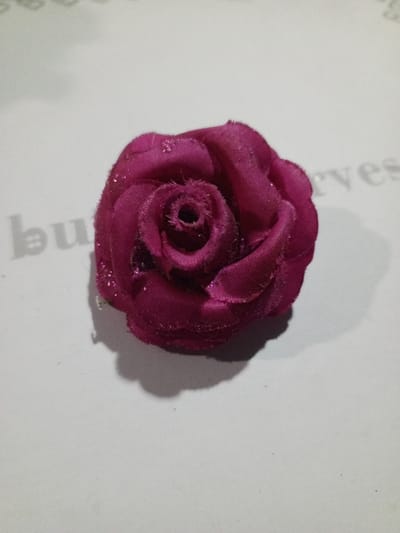 A plush, dark rose-colored flower rests on a textured surface. The rose's petals spiral inward toward a central opening, displaying a slightly fuzzy texture. The flower casts a distinct shadow on the pale backdrop. Faint text, possibly reading "bu...ves", is subtly visible beneath the flower, suggesting the backdrop might be paper or a similar material with printing on it.