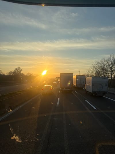 The image depicts a multi-lane highway scene at golden hour, consistent with sunset, in or near San Lazzaro di Savena, Italy. The perspective is from inside a vehicle, indicated by the partial interior frame visible at the top and dust/reflections on what appears to be a windshield. A low, intensely bright sun is positioned on the left side of the horizon, casting a strong glare and elongated shadows across the dark asphalt road surface. The sky transitions from a vibrant orange-yellow near the horizon to a lighter blue overhead, streaked with numerous thin, horizontal cloud formations.

Multiple vehicles are present in the highway lanes, traveling away from the viewer. The rightmost lane contains a series of three heavy goods vehicles (trucks), including a white box truck and a light grey semi-trailer. In the adjacent lane to the left, a silver sedan with illuminated red tail lights is visible. Additional vehicles are discernible further in the distance in the leftmost lane. The highway is bordered by bare deciduous trees on both sides, and utility lines are faintly visible on the left horizon. The road features white painted lane markings. No individuals are explicitly discernible within the frame.