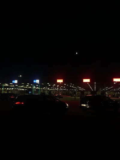 At night, a line of cars waits at a border crossing in Terespol, Poland. The border crossing is illuminated by numerous lights and illuminated signs. The sky is dark, with a sliver of the moon visible. The cars are silhouetted against the lights, and their brake lights are illuminated. The scene is calm and orderly, with no signs of conflict or distress.