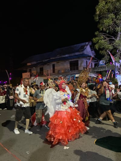 The image depicts a nighttime street scene in Kalidan Village, Kedungbanteng, Indonesia, during a parade or festive gathering. Multiple individuals are visible, predominantly dressed in elaborate costumes, moving in what appears to be a procession.

In the foreground, slightly right of center, a woman is featured wearing a red hijab, a white long-sleeved top adorned with multicolored LED string lights and small decorative items, and a voluminous tiered skirt constructed from red plastic sheeting, embellished with small white rosettes. Large white feather-like wings are attached to her back. To her immediate left, a man with short dark hair walks in a short-sleeved, collared shirt featuring a brown and white geometric pattern, dark shorts, and white sneakers; a red tassel hangs from his right pocket. Behind the winged woman, another individual wears a large, pale, mask-like headpiece with a beard and a red-and-white striped hat, a dark shirt, and light brown shorts. Further right, a person in a dark t-shirt, dark skirt, and dark shoes with rainbow-striped socks on the left leg holds an object, possibly a mobile device, raised to their right.

The background features a two-story building with a light-colored facade, dark windows, and a steep, dark roof, illuminated by artificial light. A sign, partially obscured, reads "DESA KALIDAN RT 12 RW 04." Multiple Indonesian flags are visible as decorations. String lights and other festive illuminations are present throughout the mid-ground, alongside a large tree with green foliage on the right side of the frame. The asphalt street surface shows scattered white debris. A crowd of additional participants and spectators, some wearing face masks, fills the mid-ground and background.
