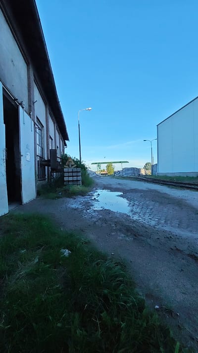 The image depicts an outdoor industrial or storage area under a clear blue sky. In the foreground, to the left, is a portion of a light-colored building with an open doorway revealing darkness within. Adjacent to the building is a pile of stacked concrete blocks. A puddle of water is visible on the wet, dirt and gravel ground in the middle of the scene, with tire tracks present. Further into the background, there are more stacks of what appear to be construction materials, possibly concrete slabs. Railroad tracks run horizontally through the midground and background. Two streetlights are present, one closer to the building and another further in the distance. A green metal structure, possibly a canopy or overhead support, is visible behind the stacks of materials. A tree with green foliage is located near the green structure. To the right, a large, plain, light-colored building dominates the background. The overall environment suggests a post-rain or humid condition. The scene is devoid of people or any visible activity. The location is identified as Goczałków, Poland.