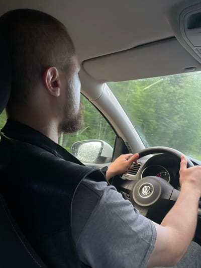 A male individual is observed from behind, driving a Volkswagen vehicle in Ореховая Гора, Russia. He has short, light brown hair and a neatly trimmed beard. He is wearing a dark, possibly leather, vest over a grey short-sleeved t-shirt. His hands are positioned on the black steering wheel, which prominently displays the Volkswagen emblem. The car's interior features a grey dashboard, air vents, and an "AIRBAG" label. Through the front windshield and the driver's side mirror, green foliage is visible, with raindrops on the glass surfaces, indicating precipitation. The passenger side sun visor is in its upright position.