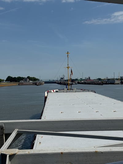 This image depicts a view from the deck of a ship or a structure adjacent to a waterway, likely in Brunsbüttel, Germany, given the location context. The scene is dominated by a large, white, flat-topped structure on the right, possibly the deck of a cargo ship, with visible rivets and some signs of wear like rust spots. To the left of this structure, a grey body of water stretches towards a distant industrial shoreline.

On the far side of the water, a line of trees separates the waterway from a cityscape featuring industrial elements, including what appear to be cranes, smokestacks, and possibly wind turbines, indicating a working port or industrial area. Several smaller vessels, including barges and what looks like a small passenger boat or tug, are visible along the shoreline. A prominent mast with a yellow top and a small flag, possibly a German flag, is positioned on the left side of the image, suggesting it is part of a ship. The water is calm, reflecting the overcast or hazy sky, which is a pale blue with wispy white clouds.

The overall impression is one of a working waterway, likely during daytime given the bright but diffused light. The perspective is from a high vantage point, looking out over the water and towards the land. There is no visible text in the image. The scene conveys a sense of industrial activity and maritime commerce in a port environment.