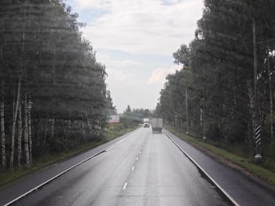 The image captures a view from a vehicle traveling on a wet, asphalt road lined with trees on both sides. The road has a double yellow line and a white dashed line in the center. In the distance, two trucks are visible, one white and one silver, traveling in the same direction. On the left side of the road, there is a billboard with text and a phone number. The sky is cloudy with patches of blue. The trees appear to be a mix of deciduous trees, possibly birch trees, given their white trunks, and evergreen trees. The scene suggests a rural or suburban setting, likely in Russia, given the mention of Melezhi. The presence of water droplets on the windshield or window through which the photo was taken is noticeable.