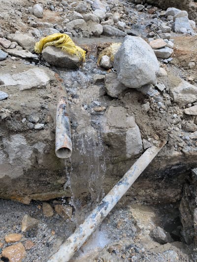 The image depicts an outdoor scene in Panamik, India, characterized by a rocky and muddy terrain. A broken pipe is visible, with water gushing out and flowing downwards. The pipe appears to be made of metal and shows signs of rust and wear. Several rocks of varying sizes are scattered around the pipe and the surrounding area. A yellow cloth is placed on top of a rock near the source of the water flow. The overall environment suggests a site where some sort of construction or repair work might be taking place, or a natural occurrence involving underground water sources. The lighting suggests it is daytime.
