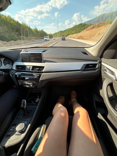 This media file captures an interior view of a car, likely a BMW based on the dashboard and steering wheel design. The camera perspective is from the passenger seat, looking forward and slightly to the right. We see a person's legs and feet in the foreground, wearing sandals and a bracelet. Sunlight casts strong shadows across the passenger's legs, indicating it is daytime.

The car is on a road, with a scenic view of a mountainous landscape and lush green vegetation visible through the windshield and side window. The road appears to be in a rural or semi-mountainous area, possibly a highway under construction given the exposed earth and machinery on the mountainside. Several cars are visible on the road ahead, suggesting moderate traffic. The sky is blue with scattered white clouds.

Notable details include the car's infotainment system, which displays a message in red and white text, along with an air vent and dashboard controls. A cylindrical object with "TEREA" visible on its packaging is placed on the car's console, possibly a product or accessory.

The overall impression is a snapshot of a journey on a sunny day, traveling through a picturesque and potentially developing region. The lighting and the scene suggest a relaxed or adventurous mood, typical of a road trip. Based on the provided location context, the scene is Prijeradi, Montenegro.