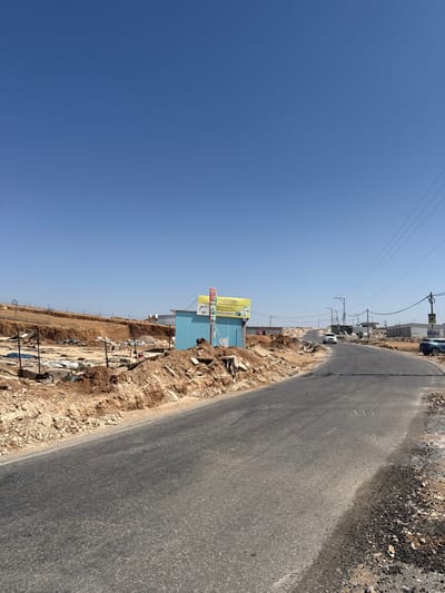 An outdoor, wide-angle image depicts an arid, unpopulated roadside scene in عناب الكبير, Palestinian Territory, under a clear, bright blue sky. A two-lane asphalt road, lacking painted markings, curves gently to the right as it ascends into the distance, occupying the lower half of the frame. The road's shoulders are composed of loose, reddish-brown dirt and gravel.

On the left side of the road, an excavated earth embankment rises, displaying layers of disturbed soil and small rocks. Immediately beyond this embankment, a light-blue corrugated metal structure stands. Attached to its front are two signs: a prominent yellow rectangular sign with green and black Arabic text and a graphic, and a smaller, partially visible red and white sign with Arabic text. To the left and behind this structure, an area of disturbed ground contains scattered debris, what appears to be remnants of a makeshift structure, and various discarded materials, indicating a possible construction site or informal settlement. A fence with wooden posts and wire is also visible in this area.

On the right side of the road, utility poles with multiple overhead power lines follow the road's path into the distance. Further down the road, a white sedan or SUV and a dark-colored SUV are parked on the right shoulder. In the far background, additional structures and buildings are visible on elevated terrain. No individuals are present within the frame. The scene is illuminated by direct sunlight, casting short shadows and indicating a clear day.