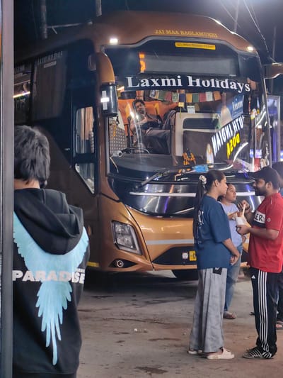 The image depicts a street scene at night, likely in Sambal, India, with a parked bus as the central element. The bus, gold and black, is identified as "Laxmi Holidays" and has "JAI MAA BAGLAMUKHI" written on its upper front. An "ALL INDIA TOURIST PERMIT" sticker is visible. Inside the bus, a man wearing glasses is seen seated, appearing to be the driver or a passenger.
In the foreground, three individuals are gathered near the bus. On the left, a person with dark hair is seen from behind, wearing a black hoodie with a large, light blue wing design and the word "PARADISE" printed below it. To the right of this person, two individuals are facing each other. A woman in a blue t-shirt and grey patterned trousers is speaking to a man in a red t-shirt and black track pants with white stripes. The man in red is holding a small white container and appears to be offering something to the woman. Another person, partially visible behind the woman in blue, is wearing a light purple t-shirt and jeans.
The environment appears to be an urban setting at night, with artificial lighting illuminating the bus and the individuals. The ground is rough and uneven, typical of a street or parking area. Several flags with Tibetan script are hanging across the front of the bus. The overall impression is a momentary pause during a journey, with people interacting before or after boarding the bus.
