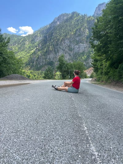 A woman with auburn hair pulled into a bun sits on a paved road, facing away from the camera. She is wearing a red t-shirt and light blue shorts, with black and white sneakers. The road is grey asphalt with white dashed lines. To the left of the road, there is a pile of dark grey gravel. Behind the woman, the road curves to the right and ascends a hill. Steep, forested mountains rise in the background, with rocky cliff faces visible on the slopes. The sky is blue with scattered white clouds. The scene appears to be in Crna Poda, Montenegro, likely on a remote mountain road.