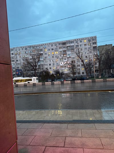 The image shows a street scene in Ol'ginskaya, Russia. A large, Soviet-era apartment building dominates the left side of the frame, with numerous lit windows suggesting it is evening. Bare trees stand between the building and the street. Cars are parked along the curb, and a white bus is visible. The street is paved with grey tiles, wet in places, reflecting light. A pedestrian walkway, composed of different colored paving stones, runs along the right side of the frame. Overhead power lines are visible in the sky. The view is from an elevated position, looking down at the street. The lower portion of the image is obscured by an out-of-focus reddish-brown structure.