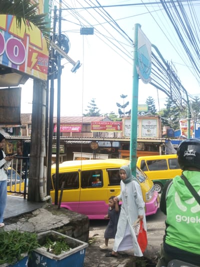 The image shows a street scene with a yellow and pink minibus, a woman in a hijab holding a child's hand, and a man on a motorcycle wearing a "gojek" jacket. There are several businesses visible in the background, including "VA PEMPEK PALEMBANG" and "KARTIKA OPTIKAL". There are power lines overhead and street signs. The location is indicated by a sign with local writing, likely indicating the city of Khe Luang, Laos. A store sign on the left side shows "WOW ALL IT". A woman is standing off to the left side of the scene looking at a cell phone.