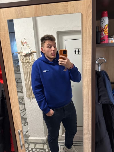 An indoor mirror selfie taken in Kaloyanovo, Bulgaria, features a young adult male positioned centrally. The male has light brown, short hair styled upwards, and is seen sticking his tongue out while holding an orange-cased smartphone in his left hand (right in reflection) to capture the image. He wears a royal blue hooded sweatshirt with a small rectangular "TOMMI" label on the chest, dark blue jeans with his right hand (left in reflection) in a pocket, and light-colored sneakers. A multi-colored braided bracelet is visible on his left wrist (right in reflection).
The mirror, framed in light wood with a dark outer border, reflects the immediate surroundings. To the left of the male's reflection, a white wall displays a macrame wall hanging below a framed card. Below these items, a patterned floor resembling a black and white mosaic and a staircase with white risers are visible. To the right of the male's reflection, a white door with black pictograms indicating male and female restrooms is present. Further right, a section of a wooden cabinet or shelving unit is reflected, showing a pink aerosol can labeled "fa dirty kiss," a dark bottle, and small jars on a top shelf. Below this shelf, a silver-colored coat hanger holds a dark jacket.