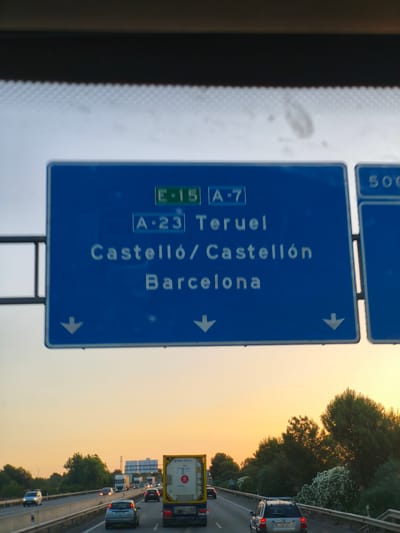 The image shows a blue highway sign with white text and downward-pointing arrows. The sign displays "E-15", "A-7", "A-23", "Teruel", "Castelló / Castellón", and "Barcelona". The sign is positioned over a highway with several vehicles, including cars and trucks, visible in the distance. The sky above the highway is a pale blue, transitioning to a warm orange as it nears the horizon, suggesting either sunrise or sunset. Trees and foliage line the side of the highway. The provided location information states that this scene is in Paterna, Spain.