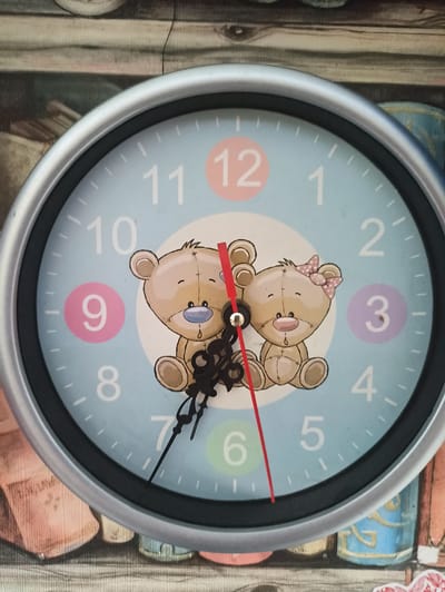The image shows a close-up of a wall clock with a light blue face. The clock has black hour and minute hands, and a red second hand. The numbers 1 through 12 are marked around the face, with some numbers highlighted in colored circles. Two cartoon teddy bears are depicted in the center of the clock face. The background of the image appears to be a shelf with books. The clock is positioned in a setting that could be a domestic or commercial interior. The photograph was taken in Promyshlennaya, Russia.