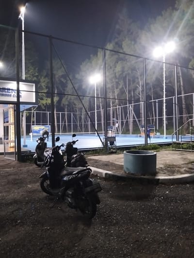 This is a night time photograph of a tennis court facility in Palohgadeng, Indonesia. The court itself is illuminated by overhead lights, and the surrounding area is dark. Several motorcycles are parked in the foreground on a paved surface. In the background, there is a chain-link fence, trees, and a building with signage. The blue tennis court is visible with nets and a few chairs. One person is visible on the court playing tennis. The overall scene suggests a sporting facility that is open for night use.
