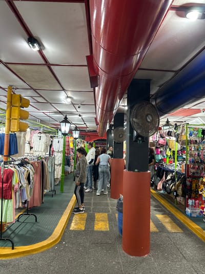 This image depicts an indoor market or covered shopping arcade located in Santa Teresita, Argentina. The scene is illuminated by artificial overhead lighting, including recessed fixtures and several green-poled streetlamp-style fixtures.

The environment features a ceiling with white panels segmented by a red grid. Large, prominent red cylindrical ducts run horizontally across the ceiling on the right side, with a blue duct partially visible further right. Below these ducts, black square support columns with reddish-orange cylindrical bases extend to the floor. At least two brown-bladed oscillating fans are mounted on these black columns.

The floor is a gray tiled or concrete surface. A pedestrian pathway is delineated by raised yellow curbs on both sides, with yellow rectangular markings resembling crosswalks visible on the floor within the pathway. A blue trash receptacle is situated next to one of the reddish-orange column bases on the right.

On the left side of the pathway, clothing racks display various garments, including light-colored tops and dresses. A large yellow multi-bulb traffic light fixture is positioned upright near the left edge of the frame. On the right side, multiple stalls present a dense array of merchandise, including handbags, colorful keychains, hats, and plush toys.

Several individuals are present. In the immediate foreground on the left, a woman with dark, curly hair, wearing a gray long-sleeved top, black leggings with white stripes, and black sneakers, stands facing right, observing the merchandise. She carries a dark fanny pack or crossbody bag. Further down the pathway, multiple other individuals are visible; one man in a white hooded sweatshirt and jeans stands facing away from the camera, and another person in a light blue top and jeans is partially visible, all appearing to browse or walk through the market.