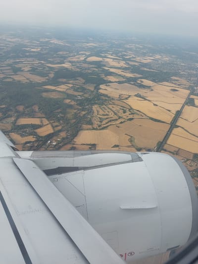 An aerial view captured from an airplane window shows a landscape of rural farmland and scattered settlements under a hazy sky. The foreground is dominated by the white, metallic casing of an airplane engine and a portion of the wing. Beyond the aircraft components, a patchwork of golden-brown fields, darker green wooded areas, and small clusters of buildings stretches towards the horizon. A road or railway line bisects the landscape in the middle distance. The image is a still, observational shot, typical of an in-flight photograph, and offers no discernible human activity or interaction. The specific geographical location within Ruscombe, United Kingdom, cannot be pinpointed solely from the visual information provided in the image.