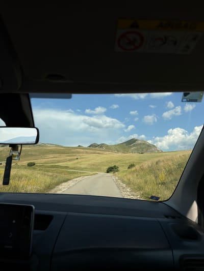 The image is taken from inside a vehicle, looking out through the windshield. The foreground shows the dashboard, with a portion of the infotainment screen visible on the left. Above the windshield, on the sun visor, an "AIRBAG" warning sticker is affixed, along with other safety symbols. A rearview mirror hangs from the windshield, reflecting a mountainous landscape.
The midground and background depict a rural, hilly landscape under a partly cloudy blue sky. A paved road curves through rolling hills covered in dry grass and scattered bushes. In the distance, a prominent, rounded mountain peak dominates the horizon. The overall scene suggests a drive through a natural, open terrain, likely in a mountainous region. The presence of the "AIRBAG" sticker indicates the location is within a vehicle that adheres to international safety standards. Boričje, Montenegro is a village located in a mountainous region, consistent with the landscape depicted in the image.