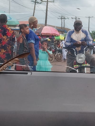 The image shows a street scene in Aketu-Oja, Nigeria. In the foreground, partially obscuring the view, is a brown feather resting on a car's windshield. Behind the windshield, a young girl in a turquoise dress is looking up and to the left, her hand near her mouth. To her left, a woman in a blue dress and head covering stands with her arms crossed, looking to the right. To the far left, a person wearing a dark patterned shirt is seen from the back, also looking to the right. In the background, a man in a blue and white track jacket and a baseball cap is on a motorcycle, holding something in his hands. Other motorcycles and people are visible further back, suggesting a busy market or thoroughfare. Several umbrellas, some colorful, are also present. The sky is overcast.