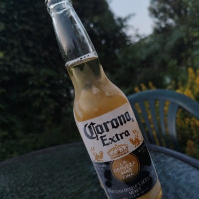 A close-up, tilted shot captures a clear glass bottle of Corona Extra beer, angled downwards. The bottle is partially filled with a pale yellow liquid, with a thin layer of foam at the top. The label features the brand name "Corona Extra" in a distinctive script, along with crowns and text in Spanish that reads "LA CERVEZA MAS FINA." Below this, further text indicates "CERVECERIA MODELO, S. DE R.L. DE C.V. MEXICO." There is a date stamp on the bottle neck that appears to read "151225 148 22:05". The bottle rests on a dark, textured surface that could be a table. In the blurred background, lush green foliage suggests an outdoor setting, possibly a garden or park. A portion of a teal-colored plastic chair is visible on the right side of the frame, also out of focus. The sky in the upper right corner is a hazy light blue, indicating it may be dusk or dawn. There are no people present in the image, and the environment appears to be serene and undisturbed. The image was taken in Luzino, Russia.
