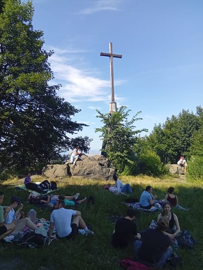 The image shows a group of people picnicking on a grassy hill near a large wooden cross. The cross stands on a rocky outcrop, with trees and a blue sky with scattered clouds in the background. Several individuals are sitting or lying on blankets on the grass, while others are seated on the rocks. Some appear to be eating or talking, while others are resting. The location is identified as Sobótka, Poland.