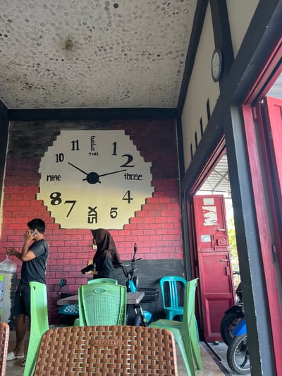 The image depicts a cafe or eatery in Welado, Indonesia. A large, painted clock face with Roman numerals and text serves as a prominent mural on a brick wall. Two individuals are present: a young man in a black t-shirt and shorts, talking on his phone while holding a plastic bag, and a woman in a brown hijab and black attire, holding a phone. Several plastic chairs, in green and brown wicker, are arranged around tables. A moped is visible to the right, and a red double door is open, revealing an outdoor area. The ceiling features a pattern of black and white circles and a single light fixture. The overall impression is of a casual, open-air establishment.