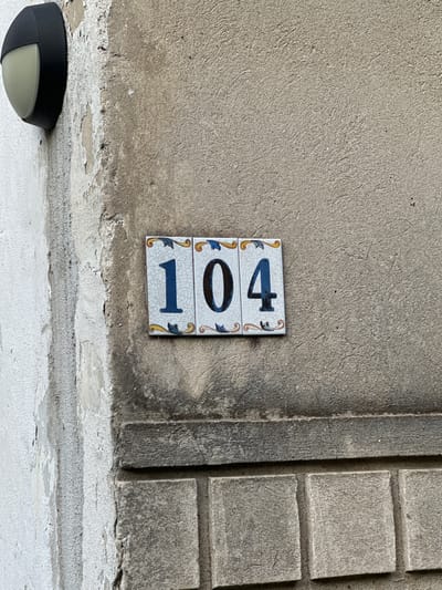 The image shows a close-up of a building facade in Lovran, Croatia, featuring house number 104. The number is displayed on a tiled plaque mounted on a textured, light brown concrete wall. The plaque consists of three square tiles, each with a blue number and decorative yellow and blue floral motifs. The leftmost tile shows the number '1', the middle tile shows '0', and the rightmost tile shows '4'. The tiles are slightly cracked and weathered, as is the surrounding concrete wall, which exhibits signs of age and wear, including peeling paint on an adjacent, lighter-colored section of the wall. To the left of the house number, a black outdoor light fixture with a rounded, translucent cover is partially visible, mounted on the lighter wall. The lower portion of the image reveals a section of the wall constructed with rectangular gray stone blocks. The overall scene depicts an exterior architectural detail of a building.