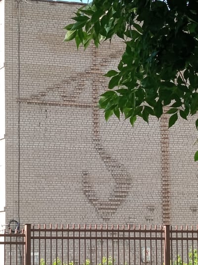 The image shows a light-colored brick building with a large swastika symbol formed by darker bricks on its facade. The symbol is partially obscured by green foliage from a tree in the foreground. A brown metal fence runs along the bottom of the image, in front of the building. The context of Novotroitsk, Russia is noted, however, no specific landmarks or identifiable features within the city are visible in the provided image. The scene is static, with no visible people or actions taking place. The lighting suggests it is daytime.